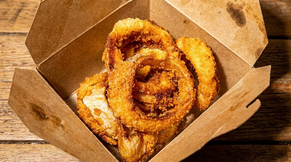 Crispy golden-brown onion rings in a brown cardboard takeout box on a wooden table.