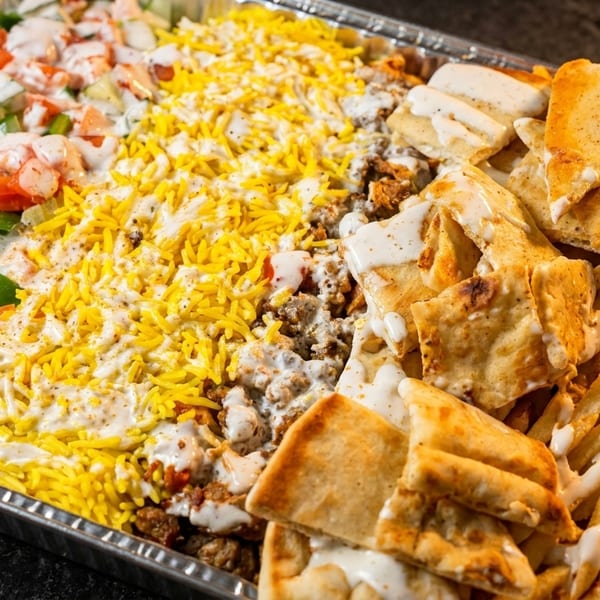 Large catering tray filled with salad, yellow rice, meat, pita bread, and french fries, all drizzled with white sauce.